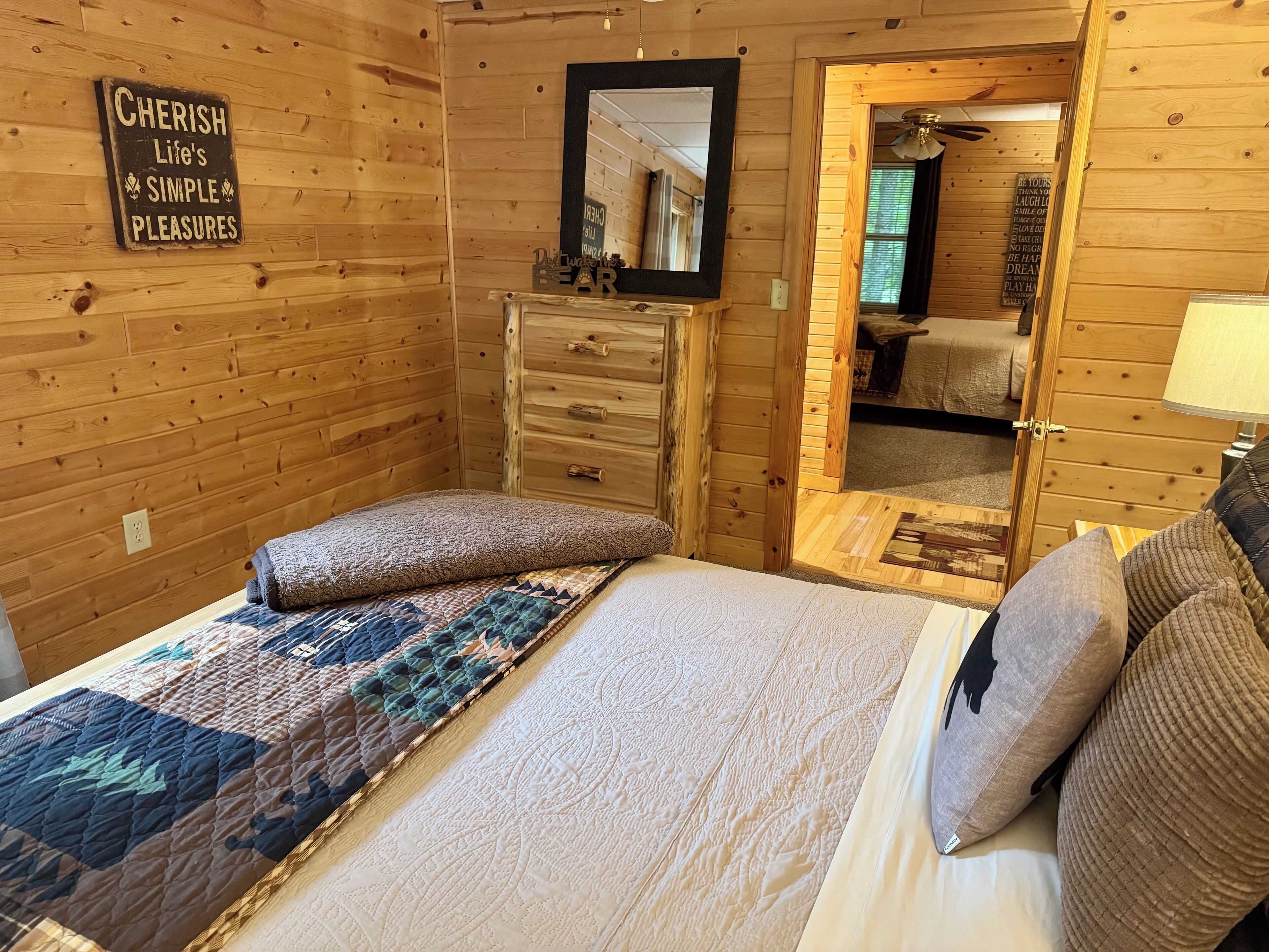 A cozy wooden room with a bed, a dresser, and a mirror. A sign on the wall reads "Cherish Life's Simple Pleasures."