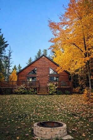 A wooden cabin with a stone fire pit in front, surrounded by autumn foliage.
