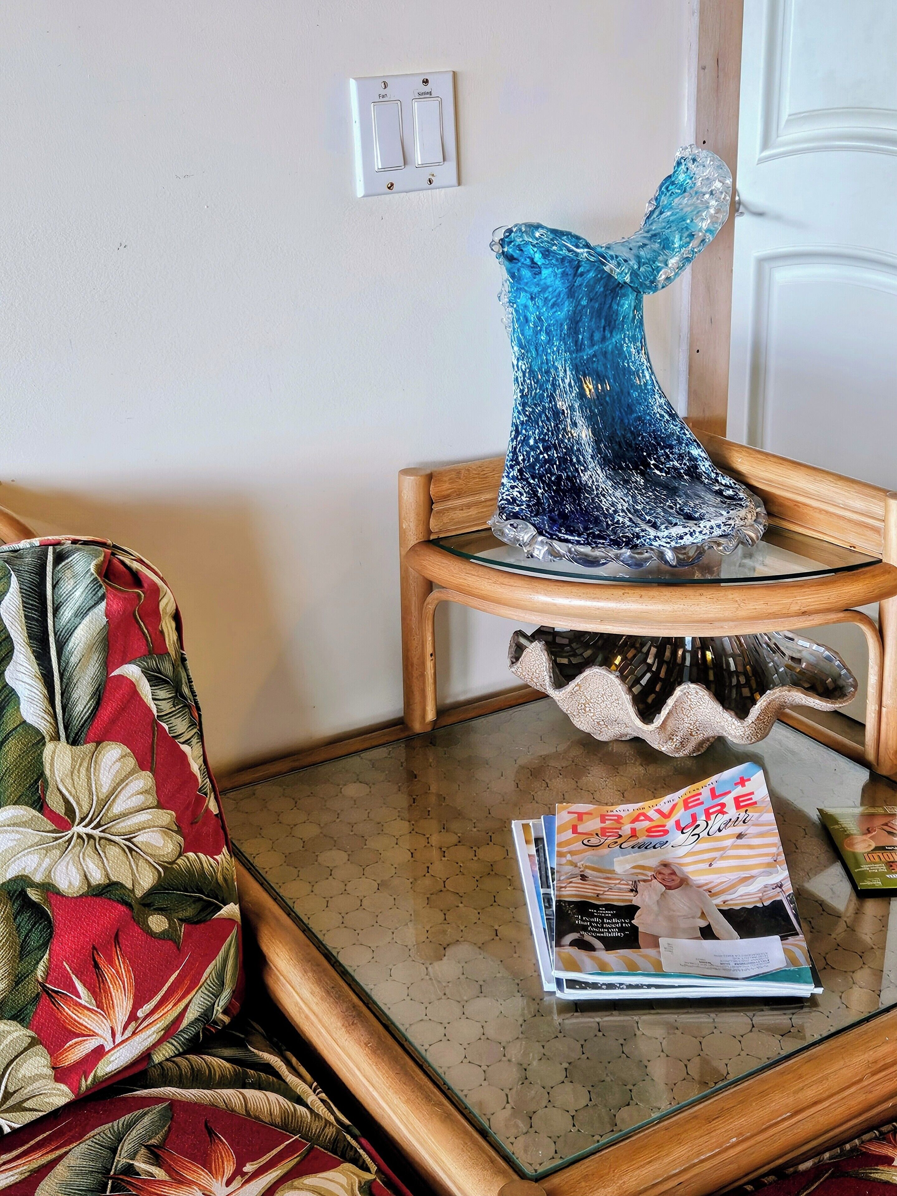 A living room with a glass coffee table, a blue decorative vase, a floral armchair, and a magazine on the table.