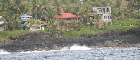 A coastal property with multiple buildings, including a house with a red roof and a larger structure with a balcony, surrounded by lush greenery and palm trees.