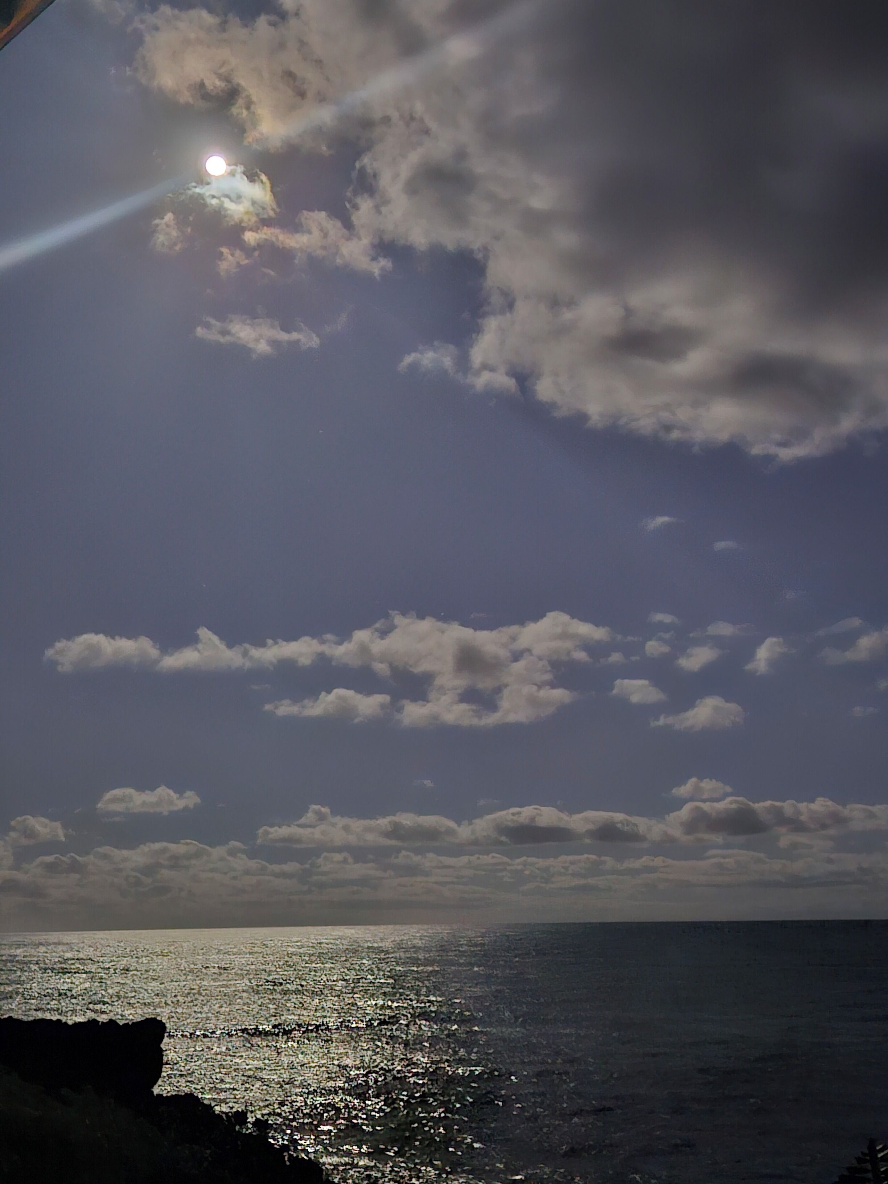 Escena costera con la luna visible en el cielo, nubes y un mar tranquilo que refleja la luz de la luna.