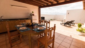 Een terras met een eettafel voor vier personen, omringd door houten stoelen. Er is een barbecue en een zithoek met loungestoelen.