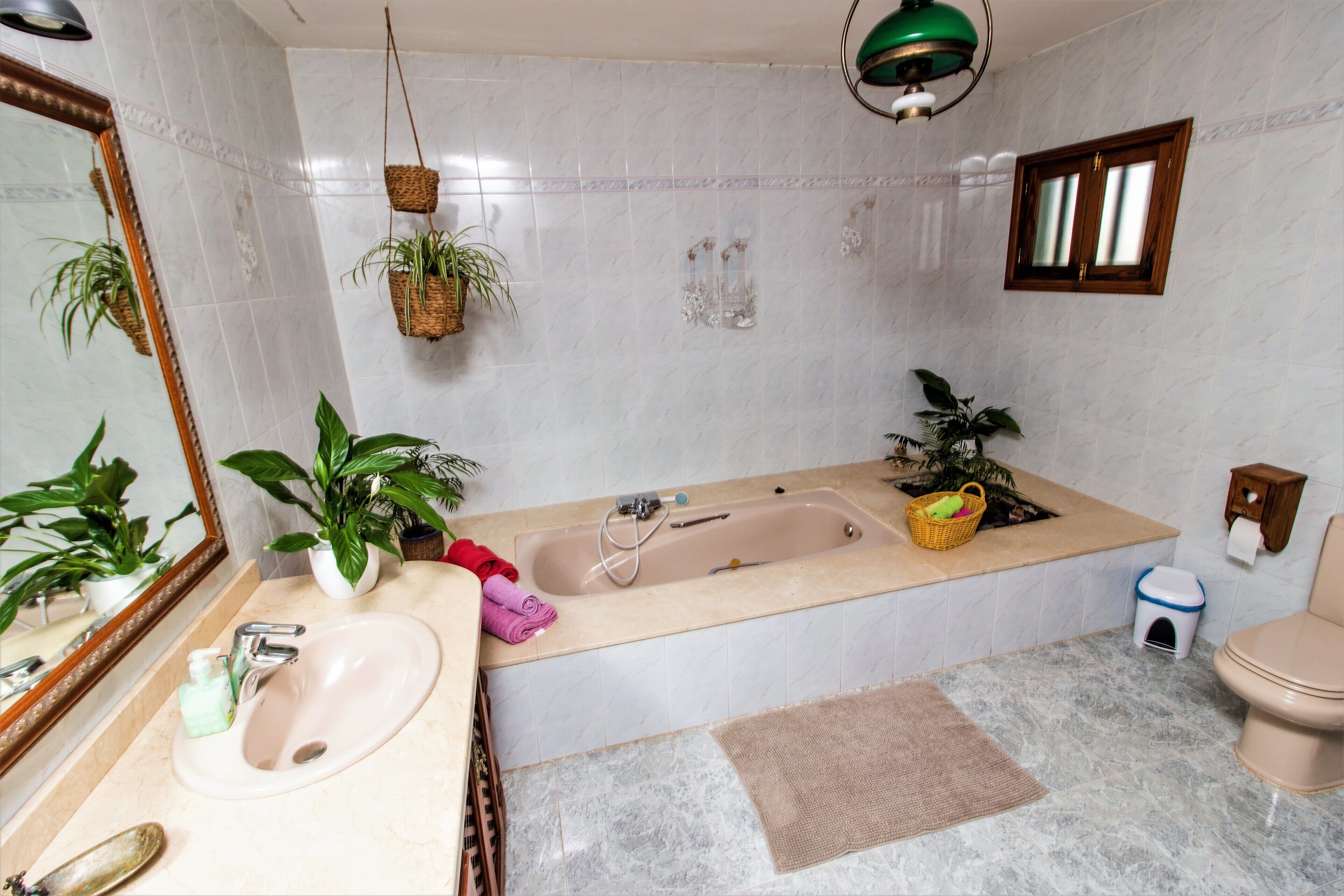 Un baño con lavamanos, espejo, bañera y retrete. Hay plantas y un perchero para toallas.