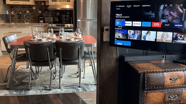 A modern kitchen with built-in appliances, a dining area with a table set for four, and a flat-screen TV mounted on a wooden cabinet displaying a streaming service interface.