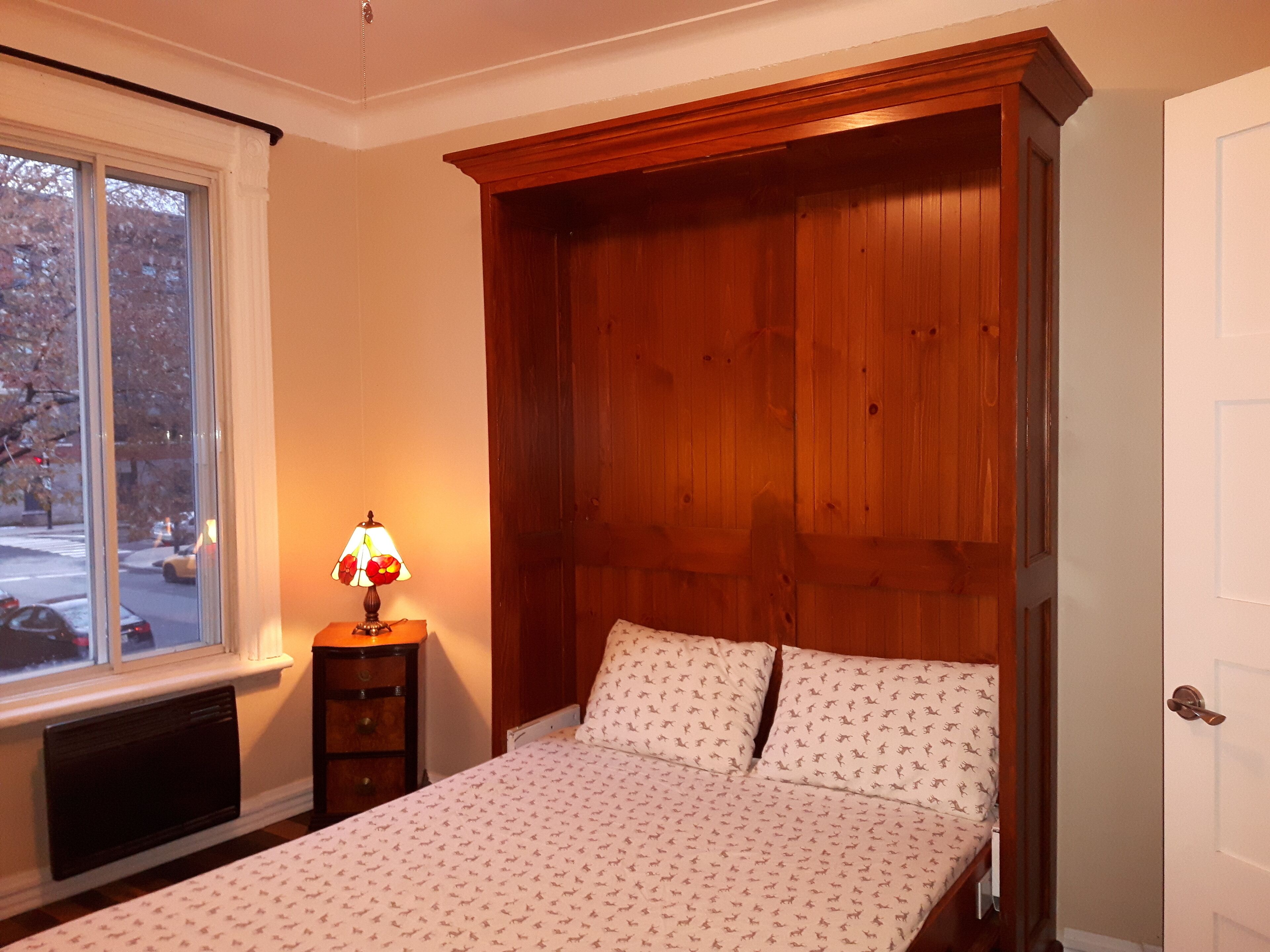 A bedroom with a wooden headboard, a bed with patterned bedding, a nightstand with a lamp, and a window with a view of the street.