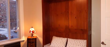 A bedroom with a wooden headboard, a bed with patterned bedding, a nightstand with a lamp, and a window with a view of the street.
