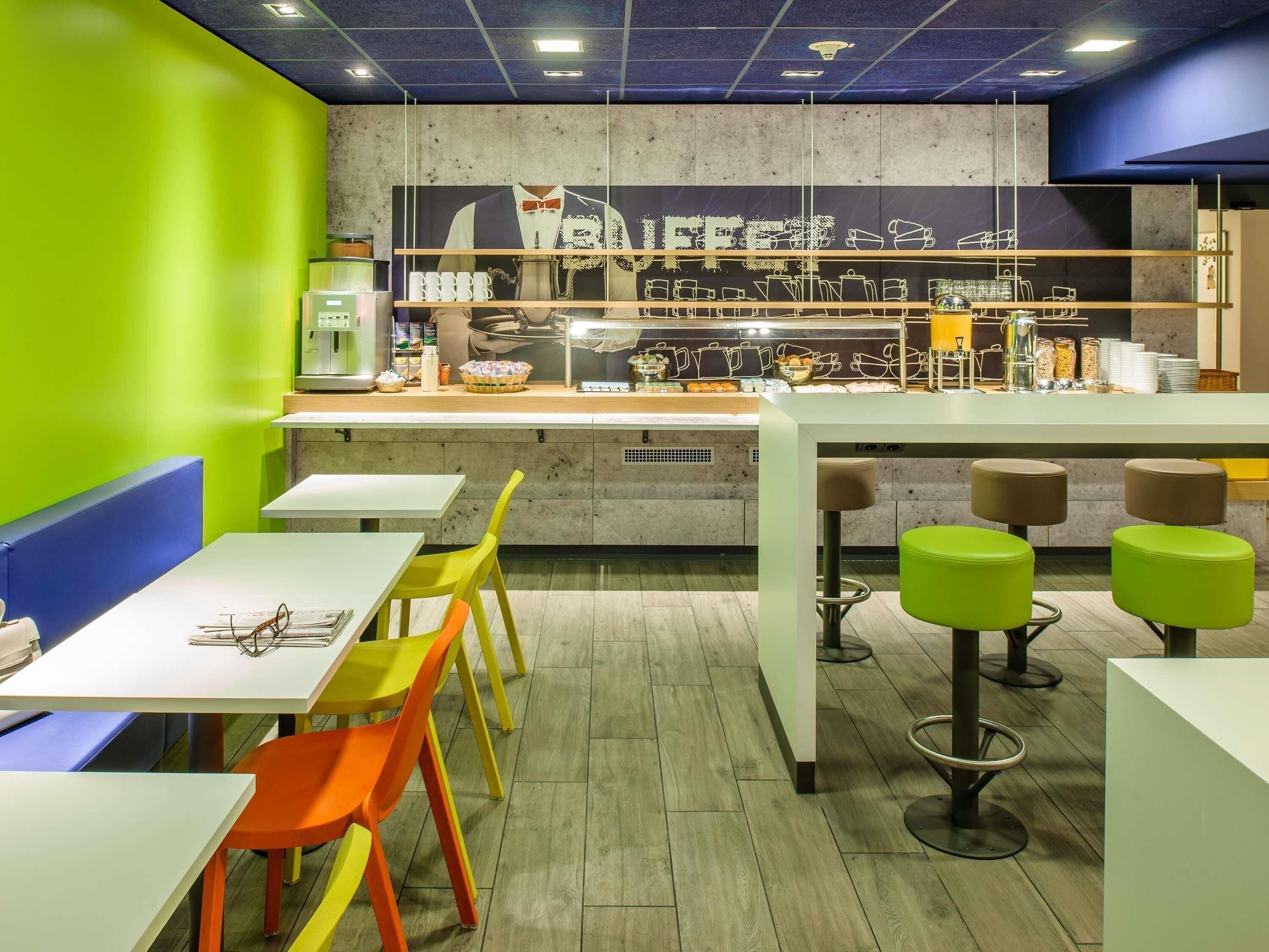 Una cafetería con un mostrador que exhibe equipo para preparar café, una zona de estar con mesas y sillas, y una pared con un mural de temática cafetera.