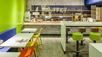 Una cafetería con un mostrador que exhibe equipo para preparar café, una zona de estar con mesas y sillas, y una pared con un mural de temática cafetera.