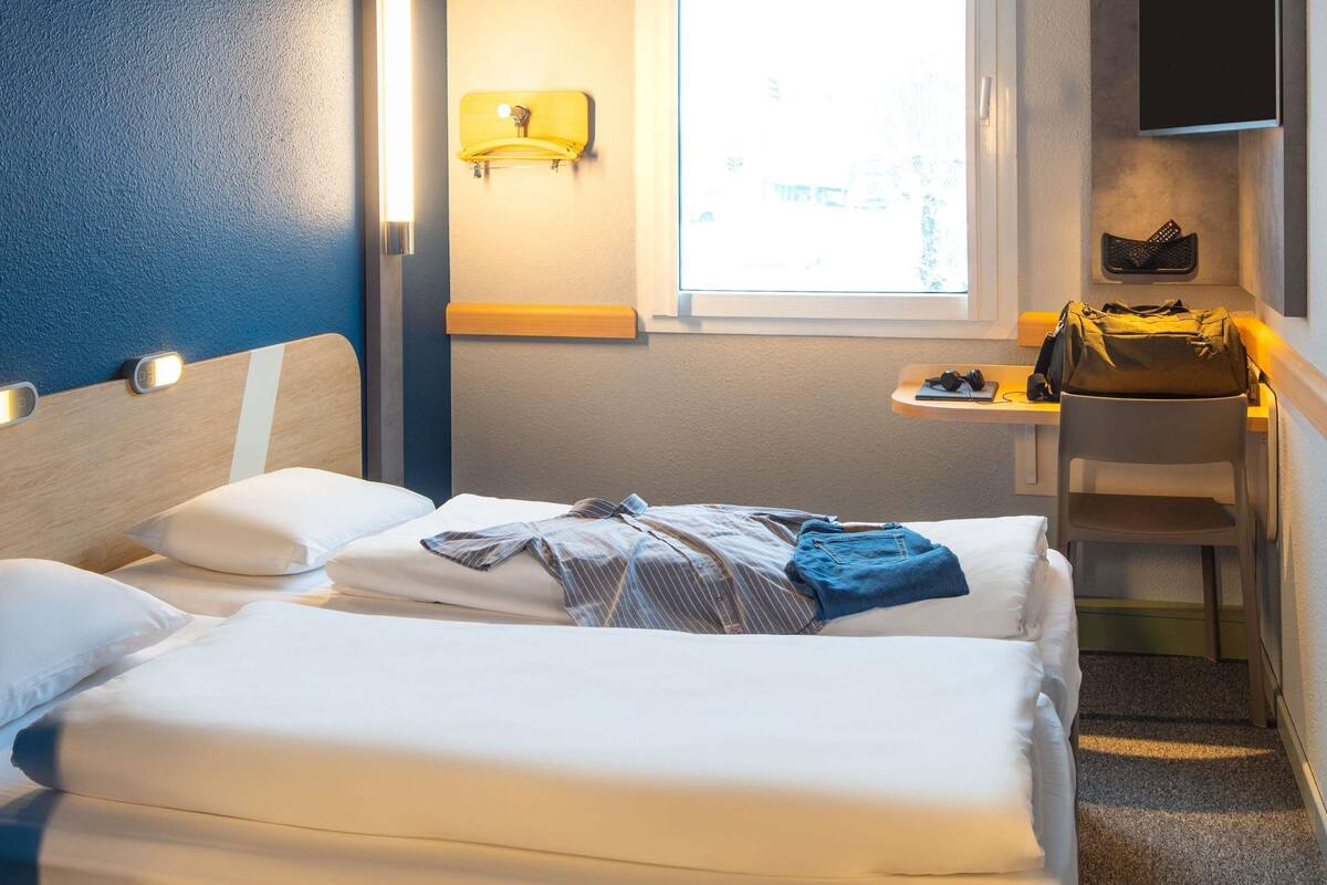 A hotel room with two beds, a small table with a bag and headphones, a window, and a wall-mounted TV.