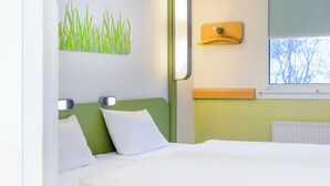A hotel room with a bed, a wall-mounted light, a window with a view, and a wall art piece. - Ibis Budget Hamburg Quickborn (Quickborn)