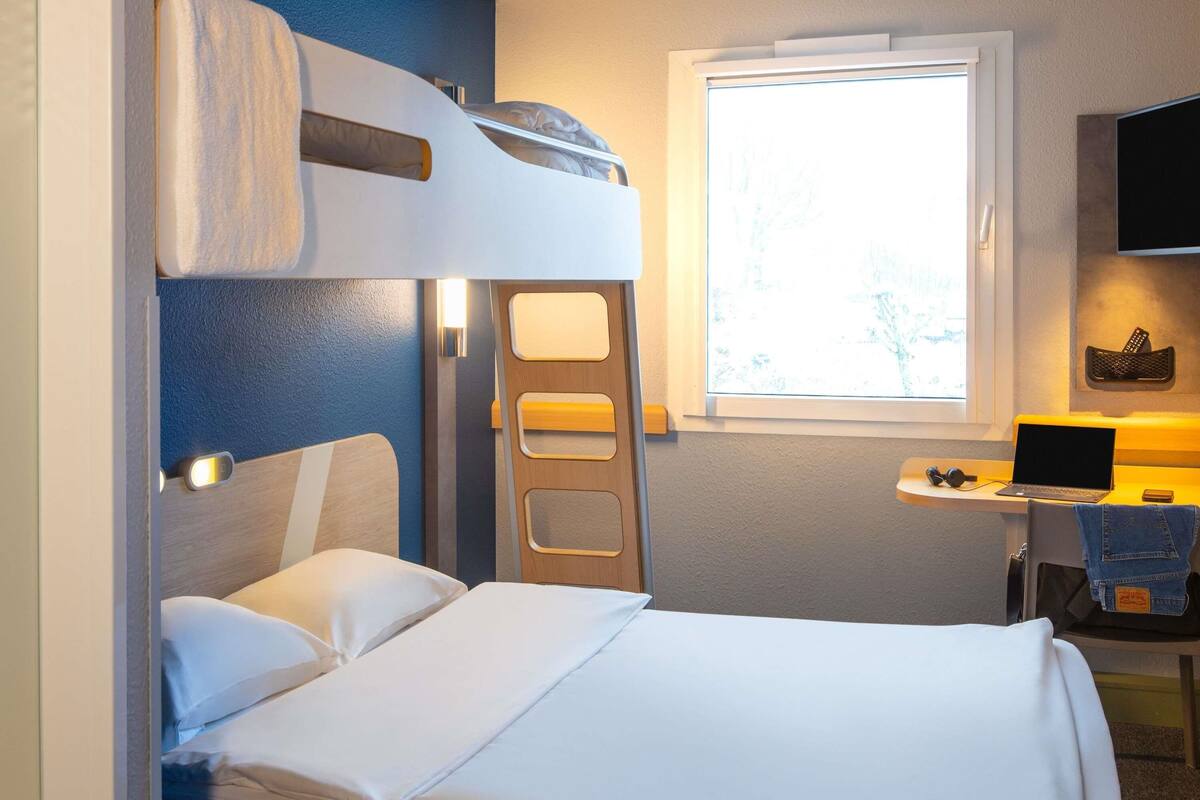 A bunk bed with a desk and a laptop, a window, and a TV on the wall.