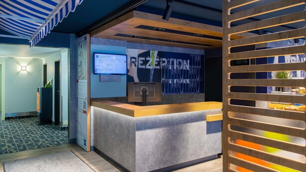 A reception desk with a computer monitor, a wooden slatted partition, and a wall with a digital display. - Ibis Budget Hamburg Quickborn (Quickborn)