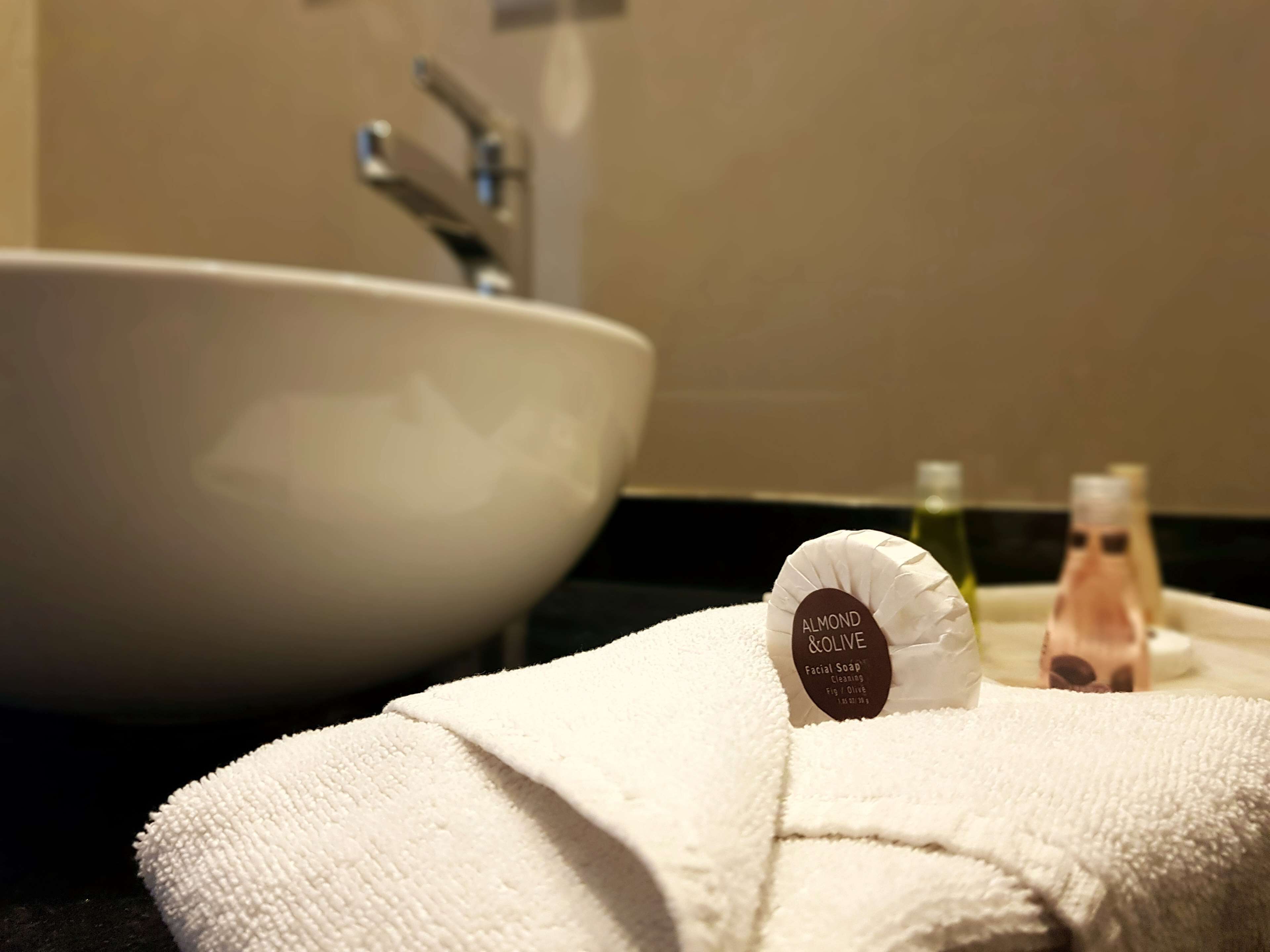 Un lavabo de baño con una pileta blanca y una toalla con la etiqueta 'ALMOND & OLIVE Facial Soap'.