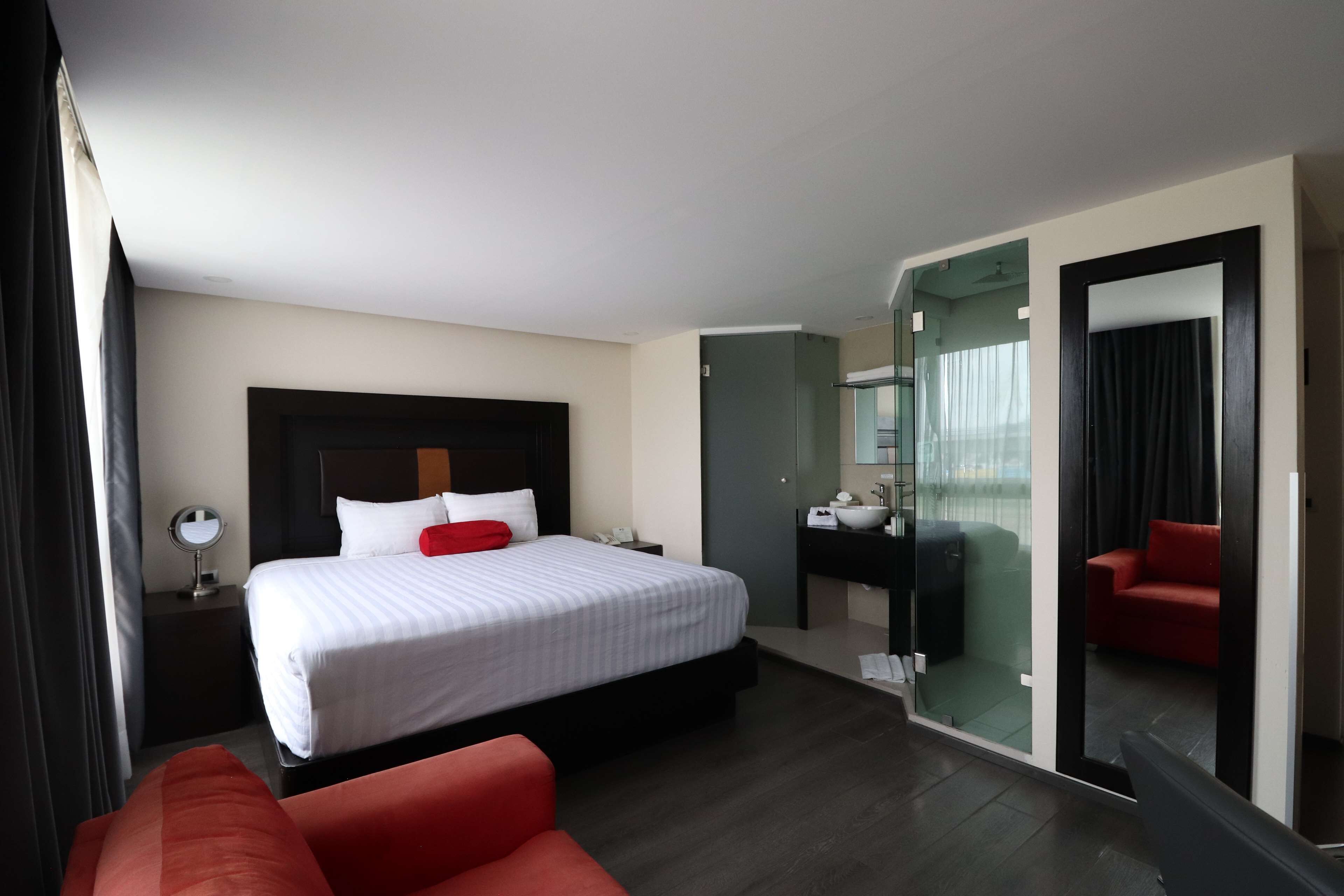 Una habitación de hotel con cama, una silla roja, un escritorio con espejo y un separador de vidrio.