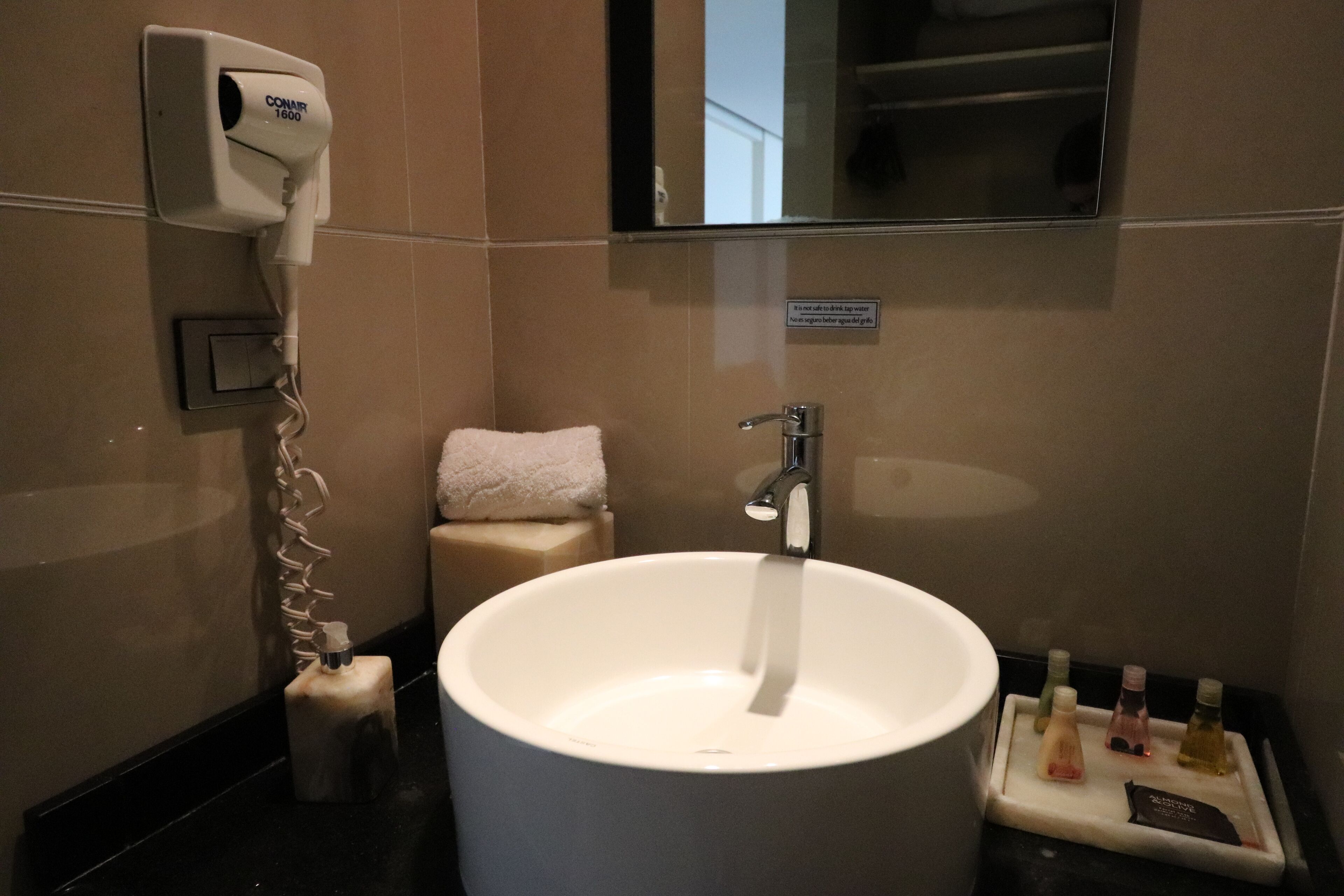 A bathroom with a large sink, a hand dryer, a towel, and a small shelf with toiletries.