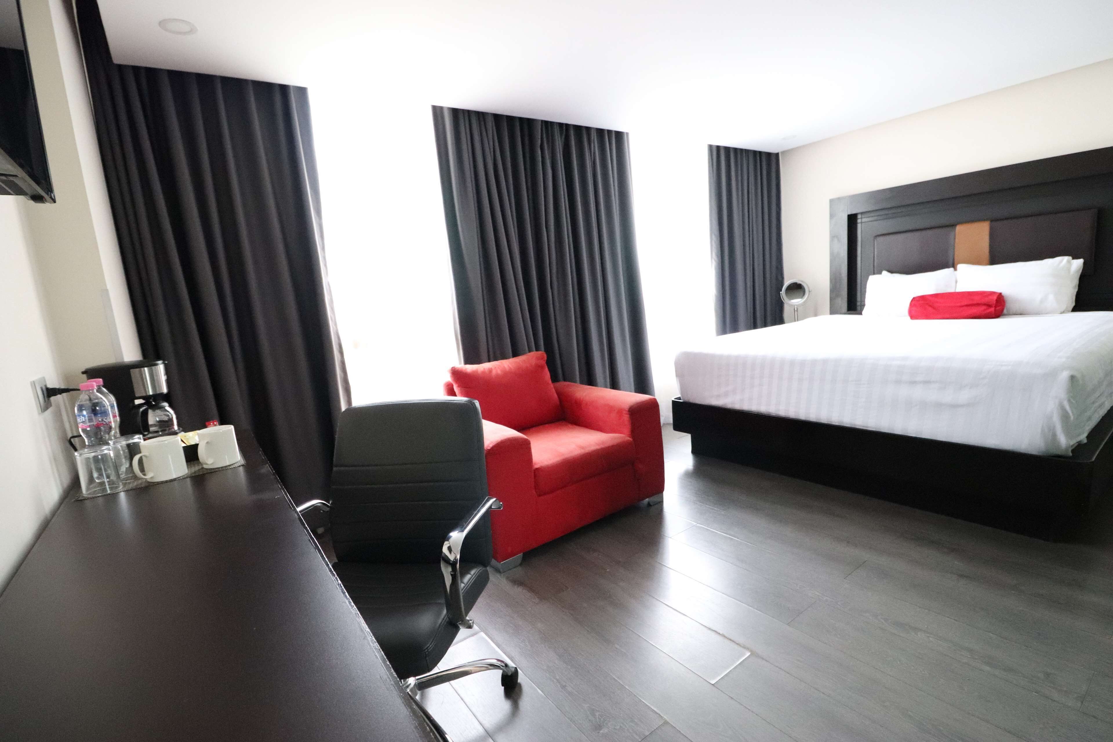 A hotel room with a bed, a desk with a chair, a red armchair, and a coffee station.