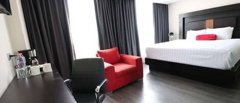 A hotel room with a bed, a desk with a chair, a red armchair, and a coffee station.