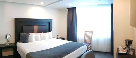 A hotel room with a large bed, a desk with a coffee maker, a chair, a mirror, and a window with curtains.