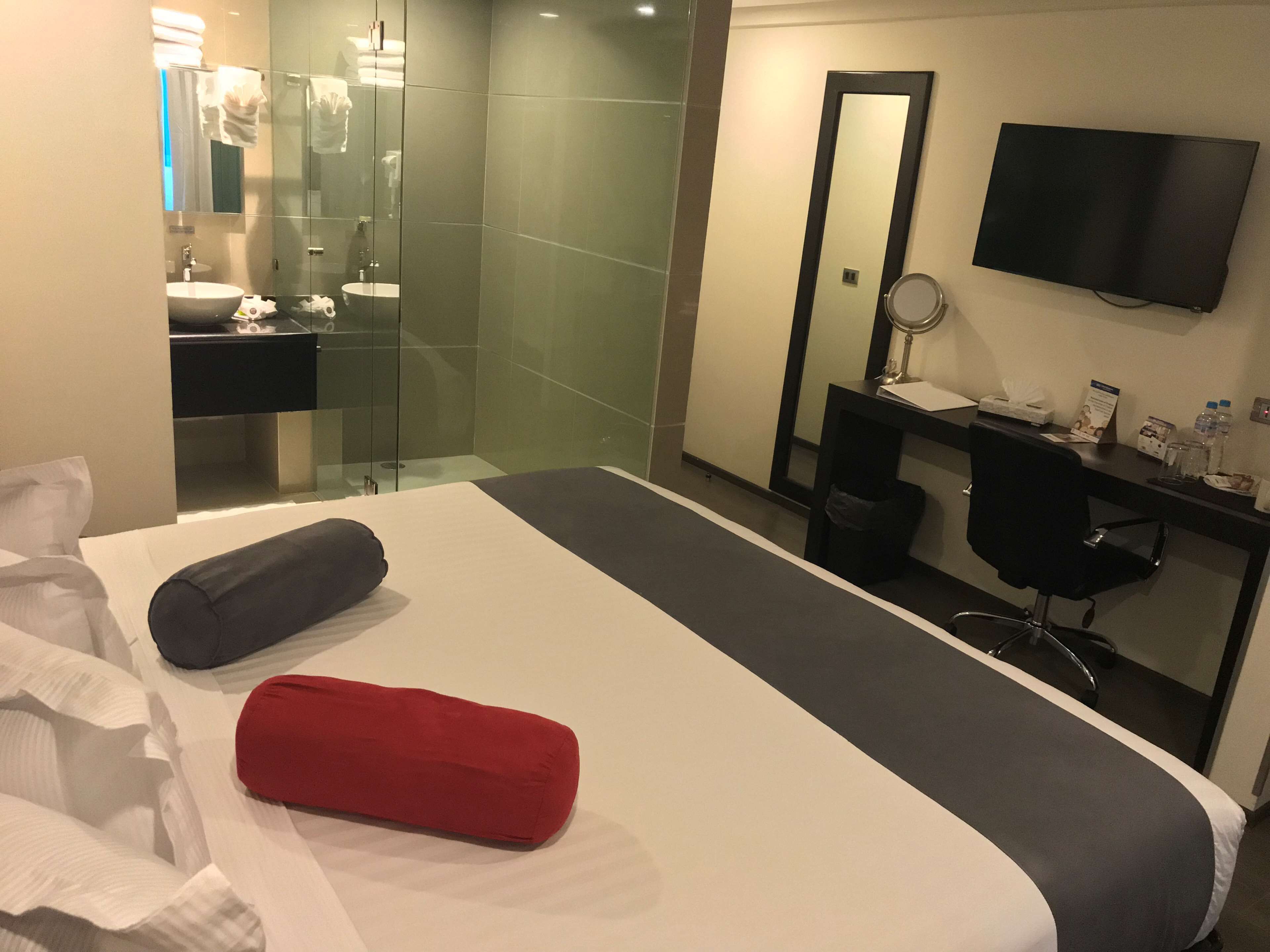 A hotel room with a large bed, a desk with a chair, a TV, a mirror, and a bathroom with a sink and glass shower.