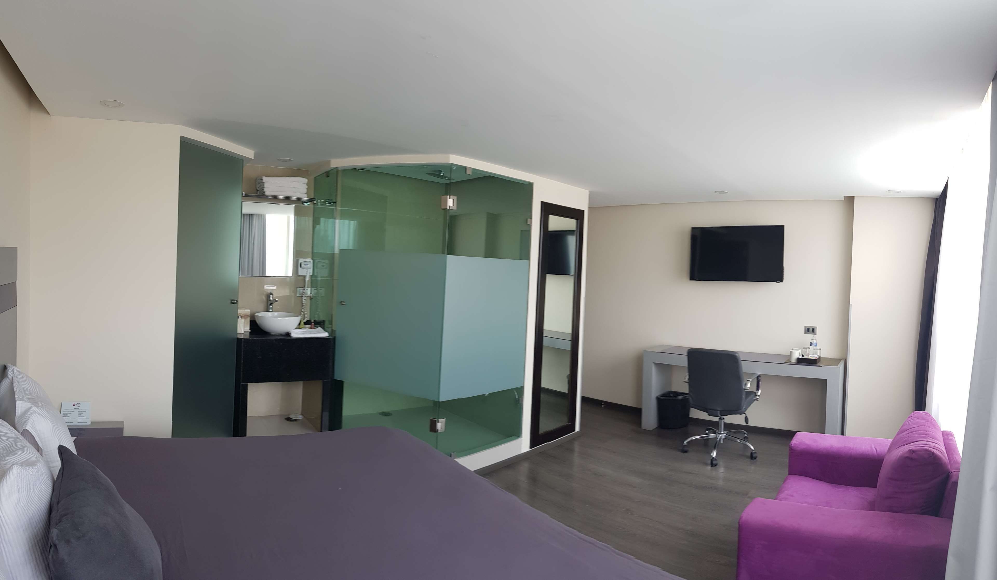 A modern hotel room with a bed, a desk, a chair, a TV, and a bathroom with a glass partition.