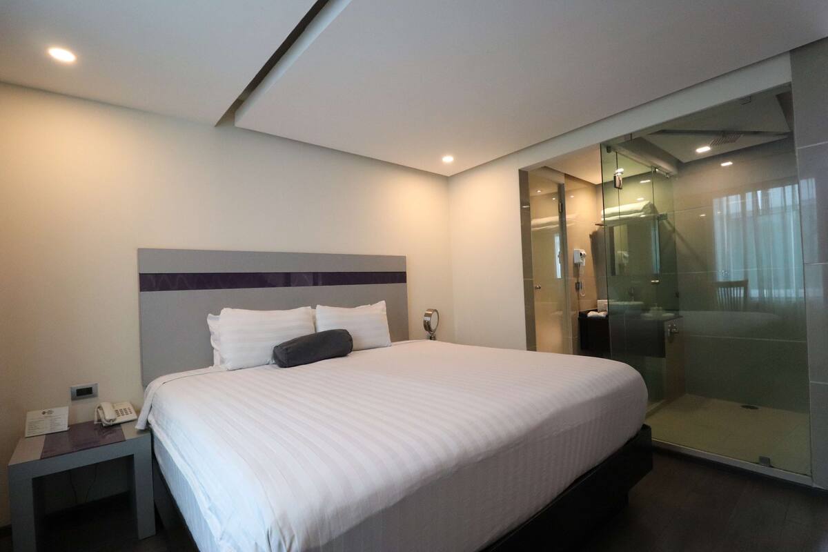 A modern hotel room with a large bed, a bedside table with a phone, and a glass-enclosed shower area.