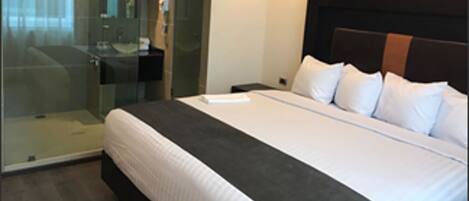 Habitación de hotel con una cama grande, una mesita de noche, cabina de ducha de vidrio y un cabecero oscuro.