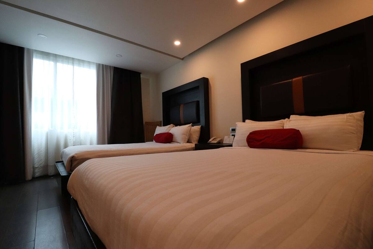 A hotel room with two beds, a large window with curtains, a dark headboard, and a bedside table with a phone.
