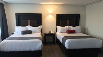 A hotel room with two beds, each with a headboard, a bedside table, and a lamp.