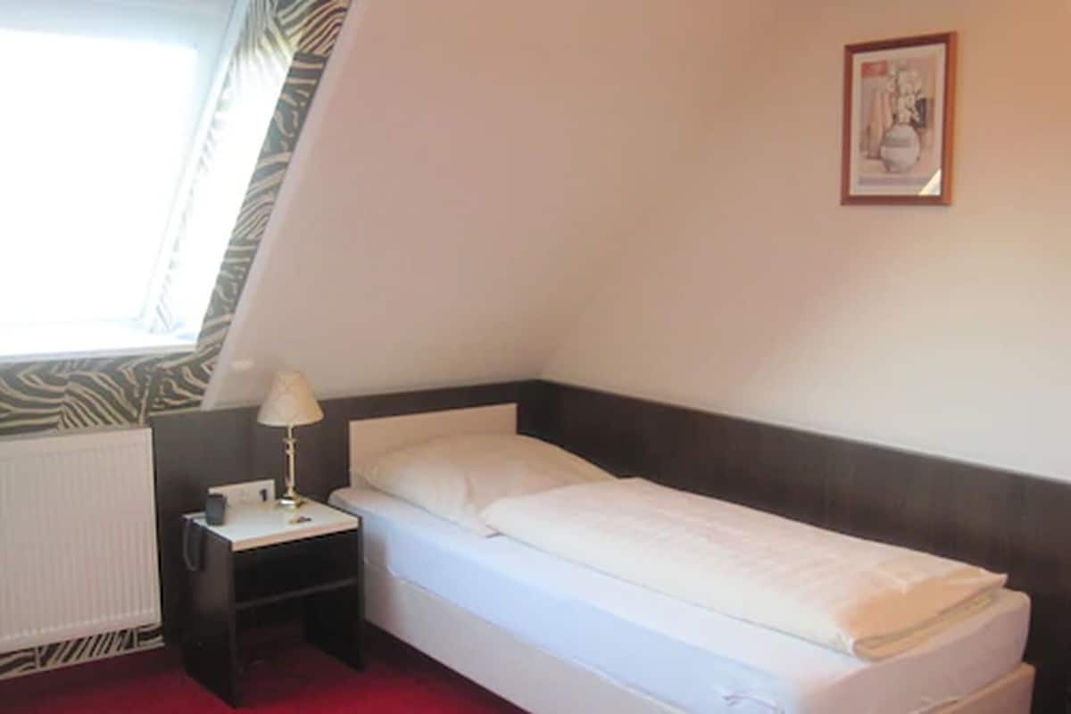 A small, single-bed room with a zebra-patterned curtain, a bedside table with a lamp, and a framed picture on the wall.