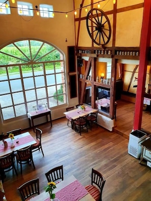 A rustic dining area with wooden tables and chairs, a large window with a view of greenery, and a wooden wheel structure. - Landgasthof Alt-Enginger Mühle (Paderborn)