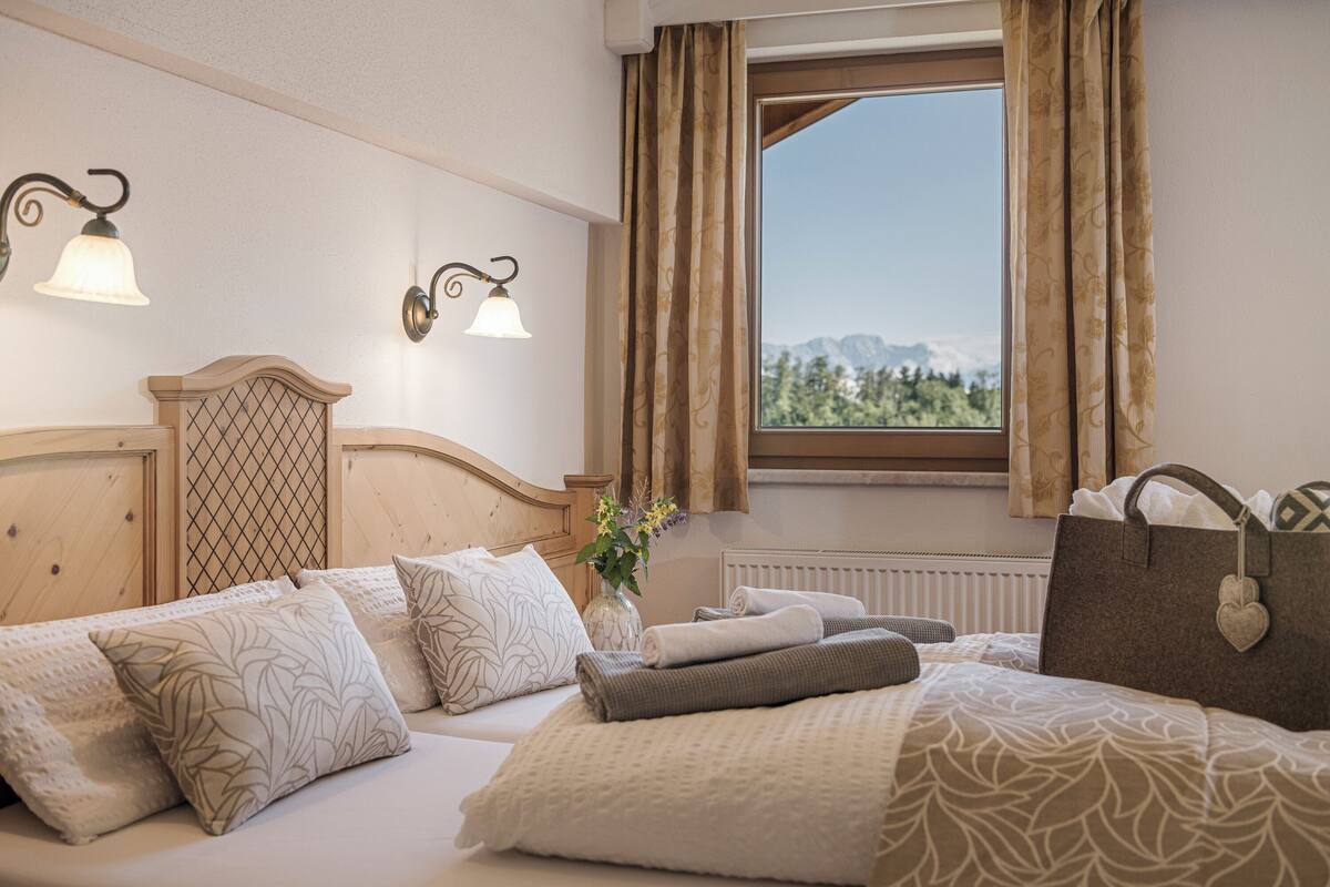 Cama bem arrumada com almofadas estampadas, um vaso com flores e vista para montanhas através de uma janela com cortinas.