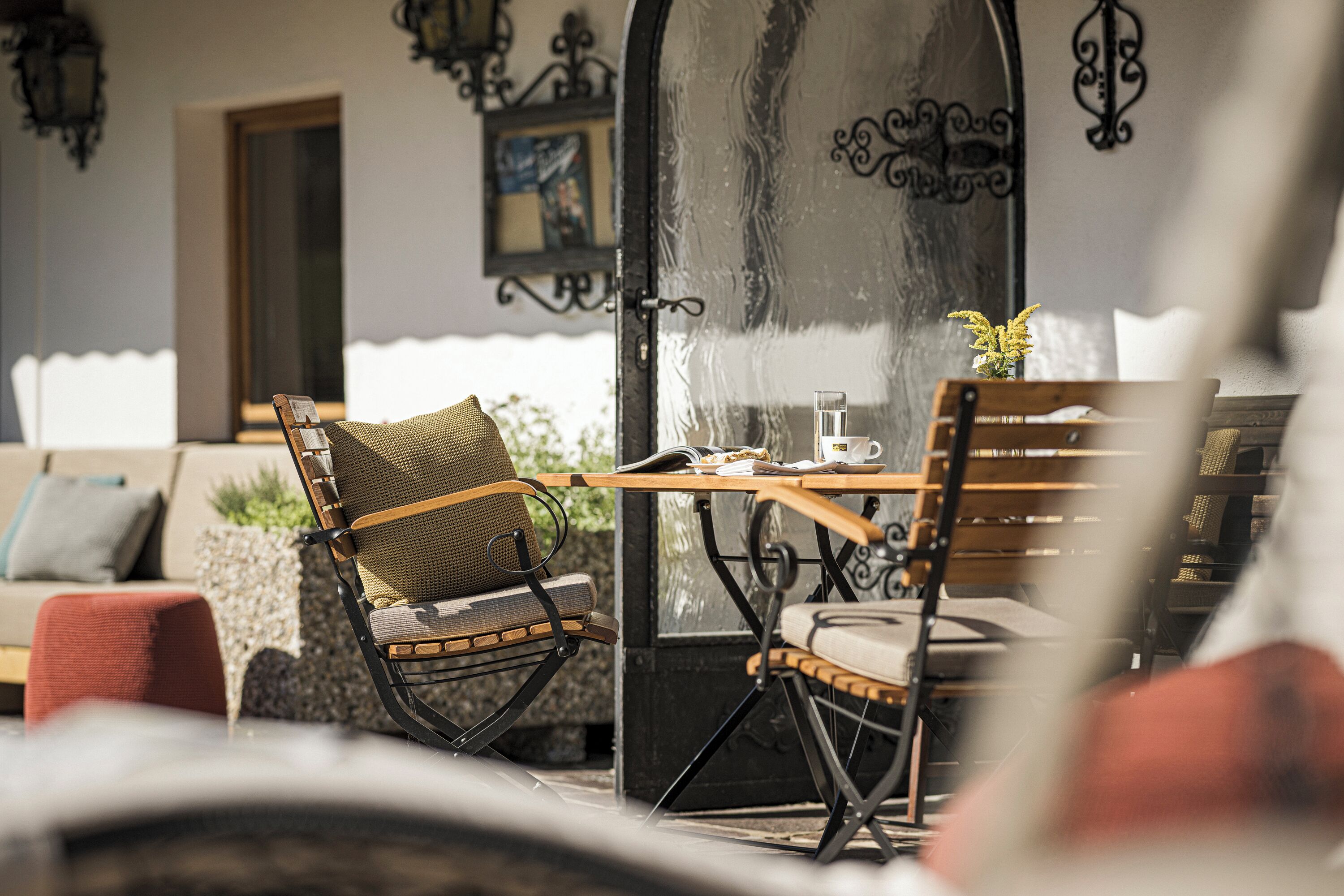 Un área de estar acogedora al aire libre con sillones de mimbre y una mesa de madera, un sofá con cojines y una pared decorativa con un espejo y obras de arte.