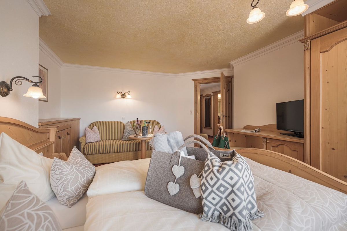 Quarto de hotel com cama, televisão, guarda-roupa de madeira e área de estar com uma cadeira e uma mesa.