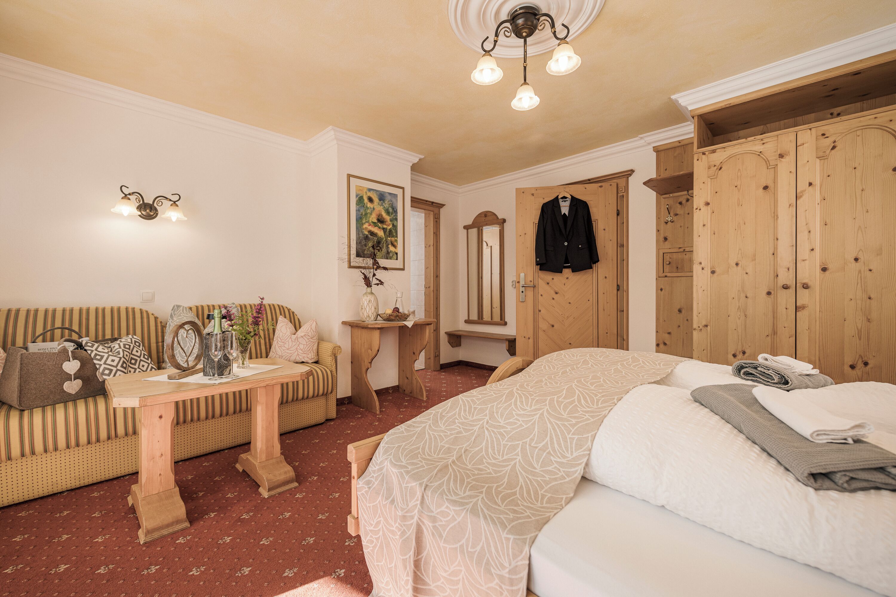 A bedroom with a bed, a wooden wardrobe, a wooden table, a sofa, and a mirror.