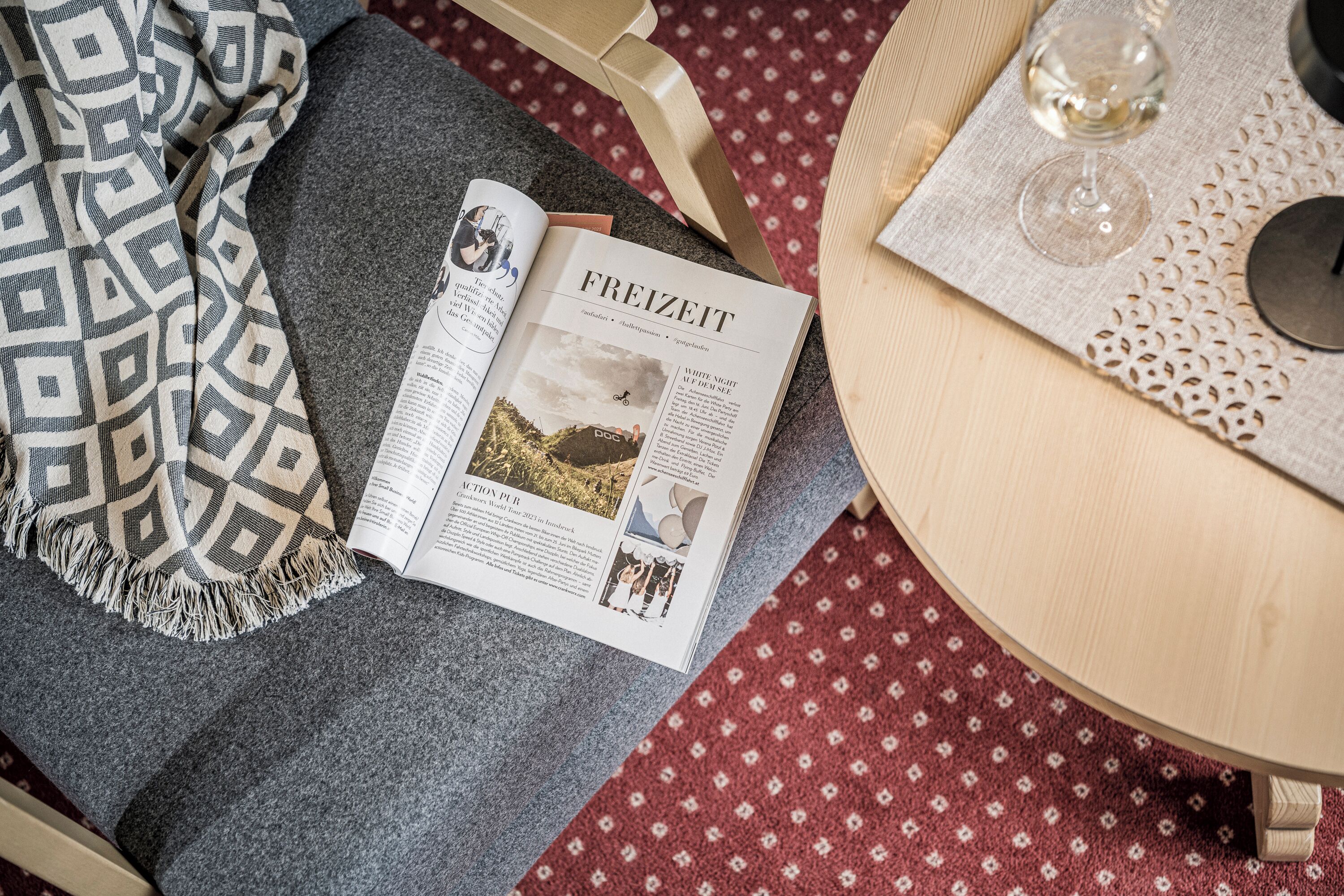Una revista abierta sobre una mesa, con una manta estampada y una copa de vino.