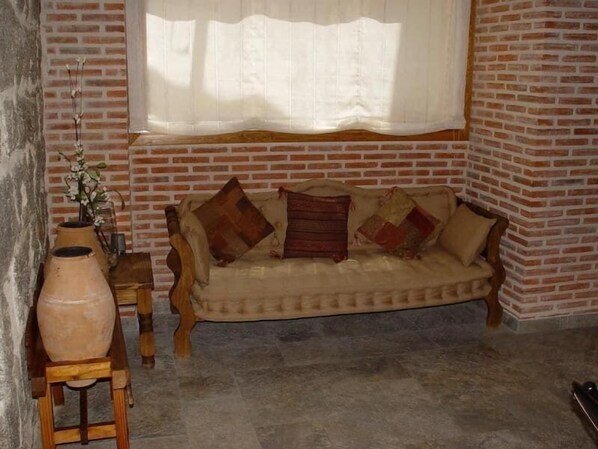 A cozy room with a brick wall, a cushioned sofa with patterned cushions, a wooden side table, and a large window with sheer curtains. - El Rastro (Avila)