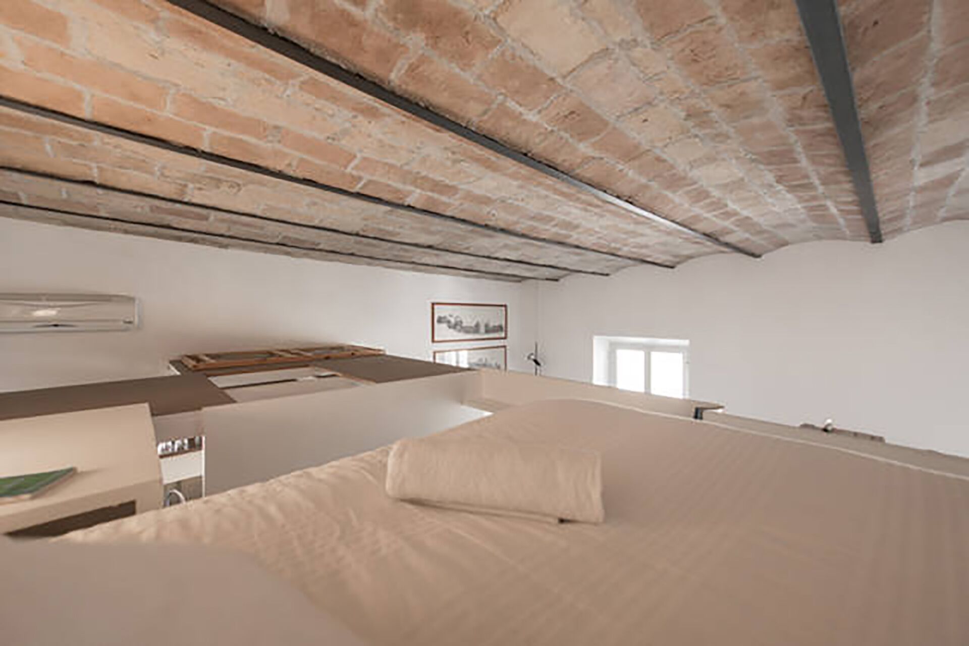 Una habitación con techo alto, ladrillo visto, una cama con almohada, un escritorio y un cuadro en la pared.