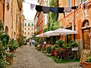 A narrow cobblestone street with buildings on either side, outdoor seating, and a bicycle. - Trastevere, Roma: design and charme (Roma)