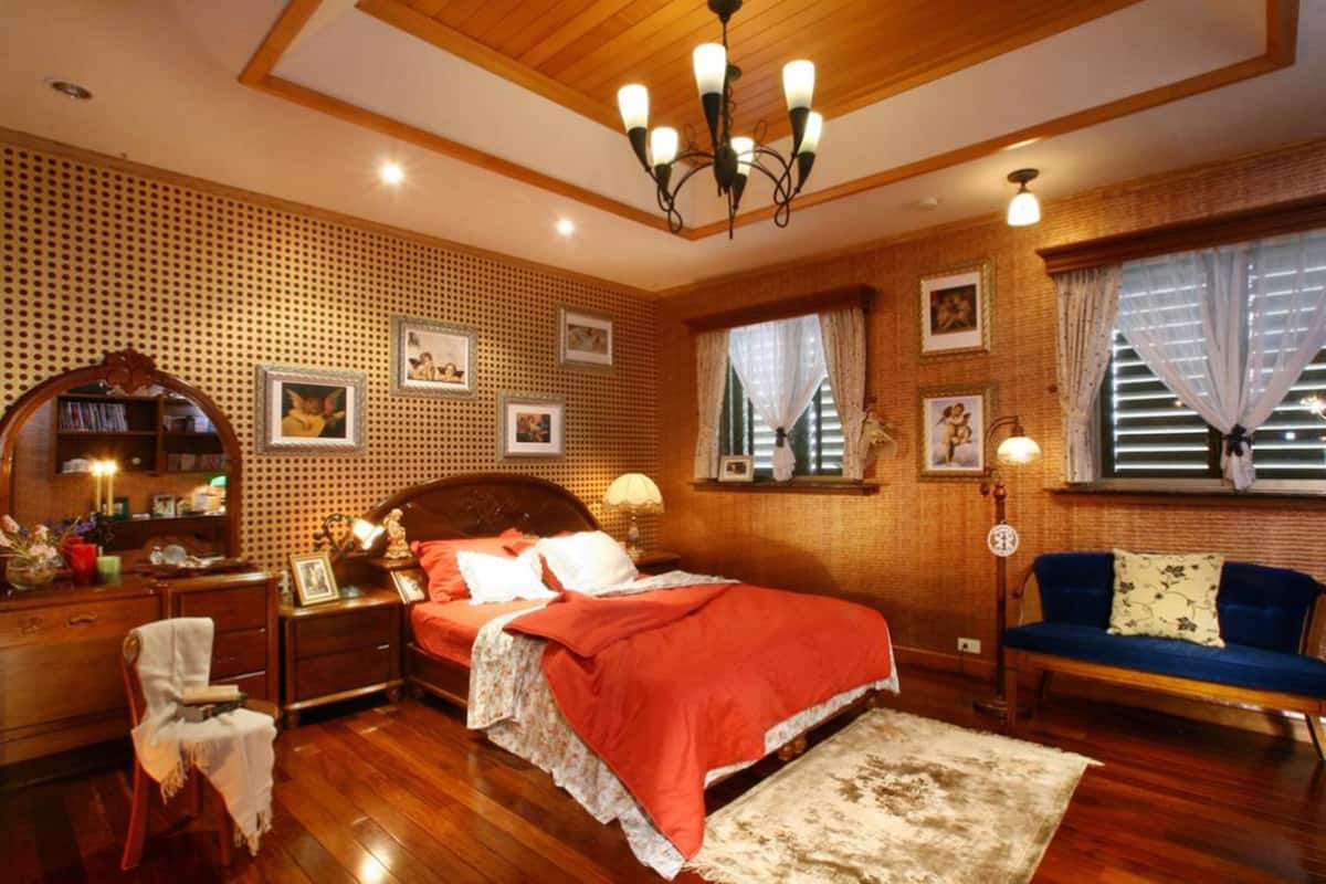 A bedroom with a wooden floor, a bed with red bedding, a blue sofa, a wooden dresser, a mirror, and a chandelier.
