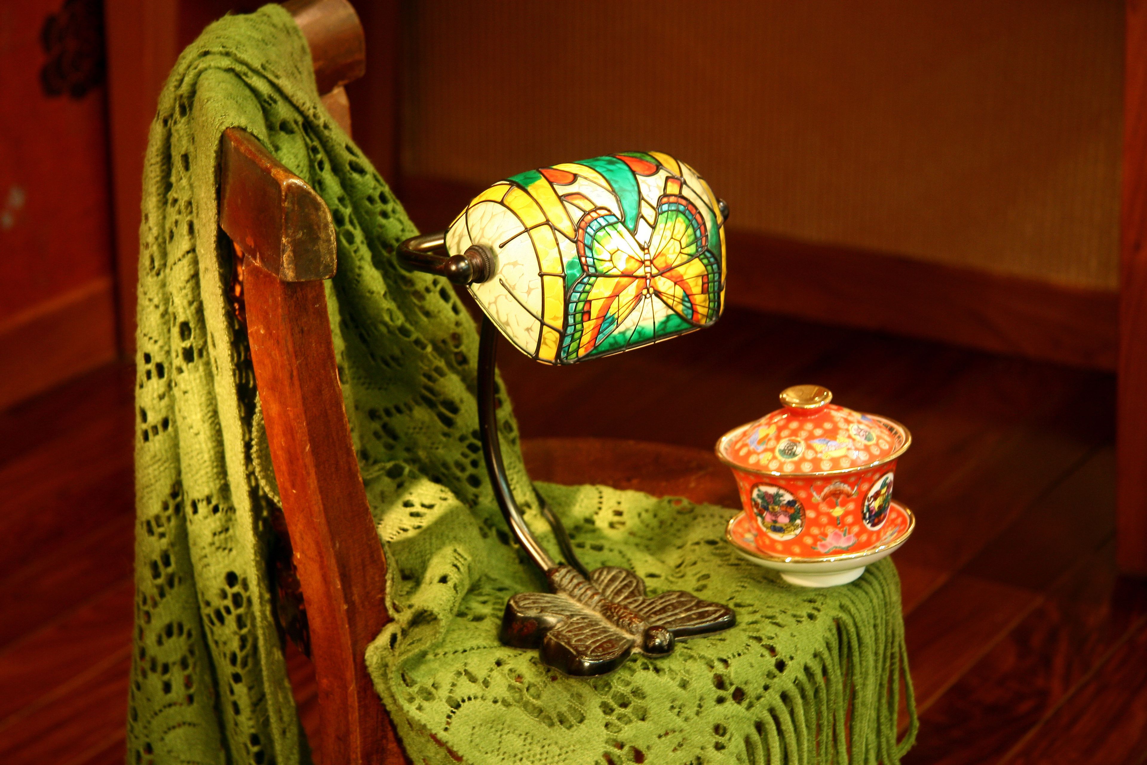 A wooden chair with a green crocheted seat and backrest, a decorative metal butterfly, and a colorful stained glass lamp.