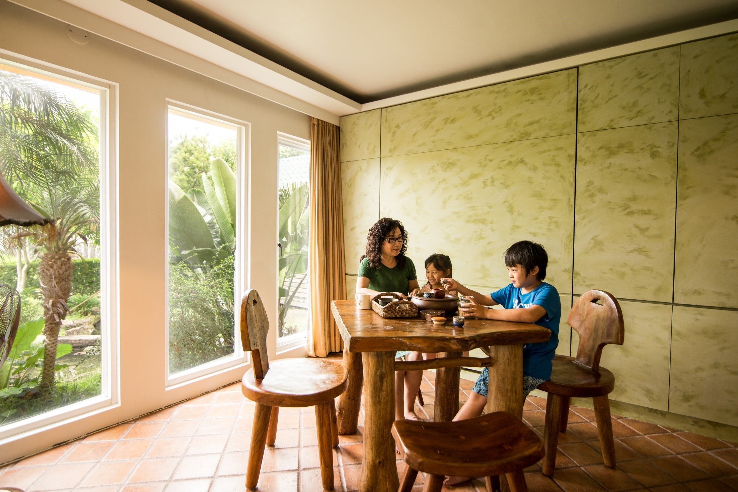 A dining area with a wooden table and chairs, large windows with a view of greenery, and a tiled floor.