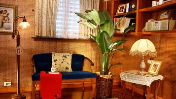 Uma sala de estar aconchegante com um sofá azul, um cobertor vermelho, uma planta grande, uma mesa com abajur e uma estante com diversos objetos.