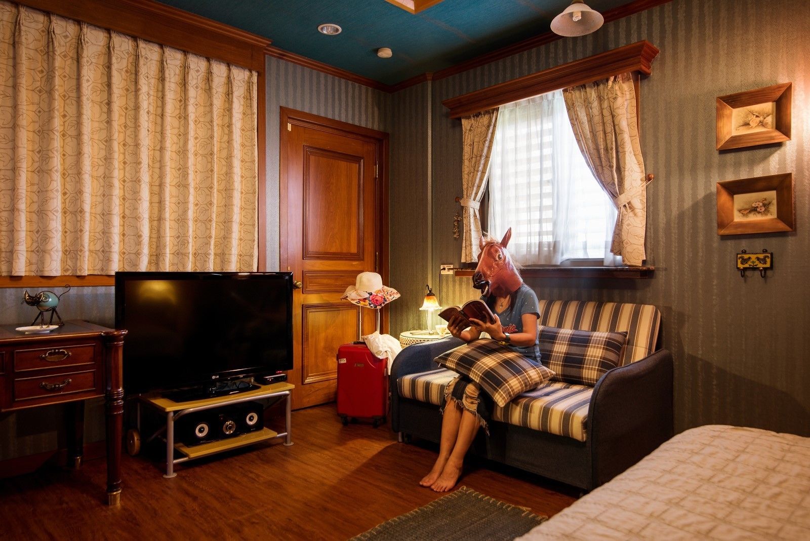 A cozy room with a television, a red suitcase, a striped sofa, and a person reading a book.