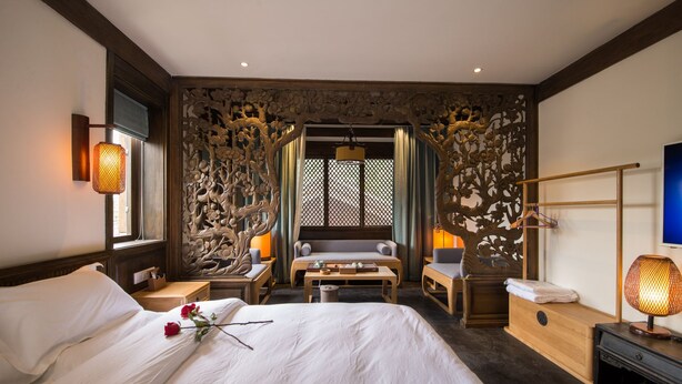 A hotel room with a large bed, a sofa, a desk, and a TV. There are decorative wooden panels on the walls and a window with a view. - Lijiang Zhuyun boutique Hotel (Lijiang)