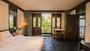 A bedroom with a large bed, a bedside table with a lamp, a small table with chairs, and a balcony with a view of greenery. - Lijiang Zhuyun boutique Hotel (Lijiang)