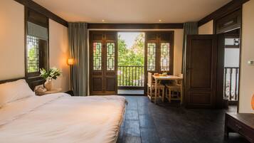 A bedroom with a large bed, a bedside table with a lamp, a small table with chairs, and a balcony with a view of greenery.