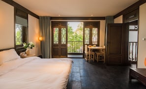 A bedroom with a large bed, a bedside table with a lamp, a small table with chairs, and a balcony with a view of greenery. - Lijiang Zhuyun boutique Hotel (Lijiang)