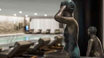 A bronze sculpture of a seated figure in a modern indoor setting with a pool and lounge chairs in the background.