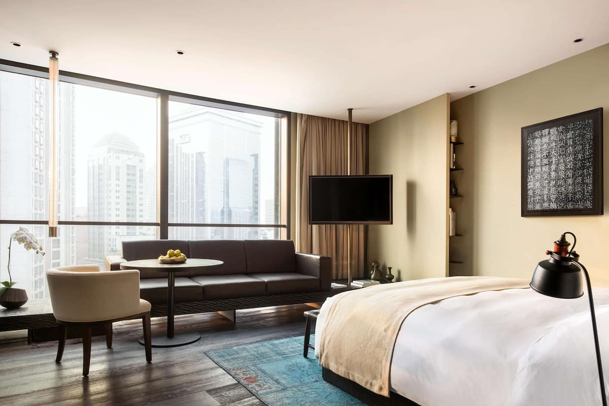 Ein Hotelzimmer mit einem großen Bett, einer Couch, einem kleinen Tisch mit einer Obstschale, einem Fernseher und Blick auf hohe Gebäude.