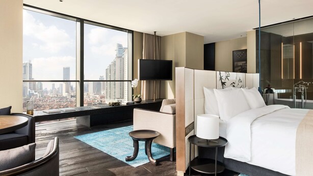 A modern hotel room with a large window offering a city view, a bed with white linens, a small side table, and a seating area with a chair and a small table.